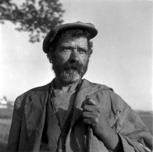 Legenda: Agricultor apoiado no cabo de uma enxada. Concelho de Almada, d&eacute;cada de 1950.